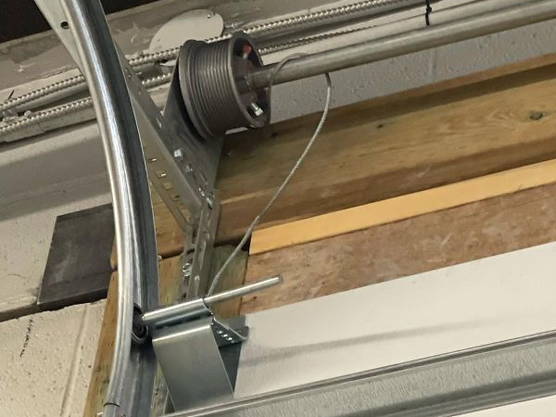 Top corner of a garage door showing the grooved cable drum on the torsion shaft, the curved track behind it and the lift cable dropping to the bottom bracket