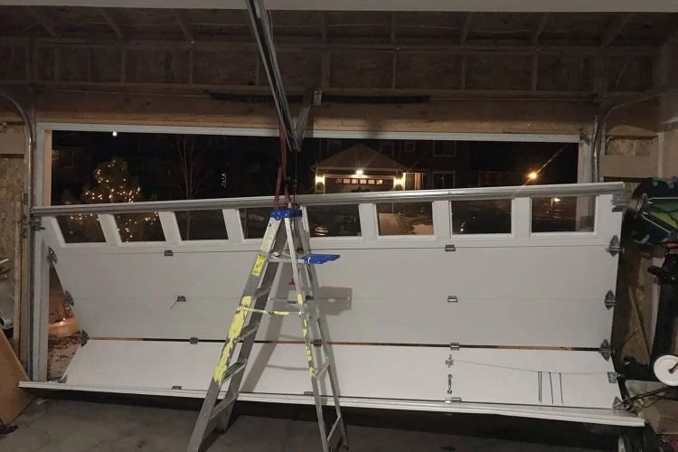 Night call in progress: a sectional door lifted clear of its tracks and propped on a stepladder inside the garage, with the empty opening and the dark street beyond