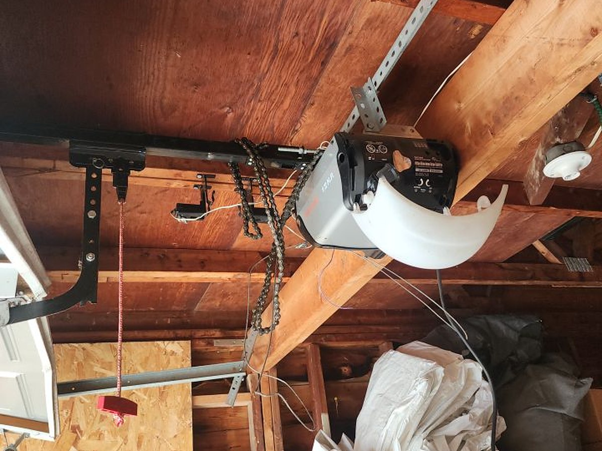 Chain-drive operator bolted to the exposed rafters of an unfinished garage ceiling, with the rail, the chain and the red manual release cord all in frame