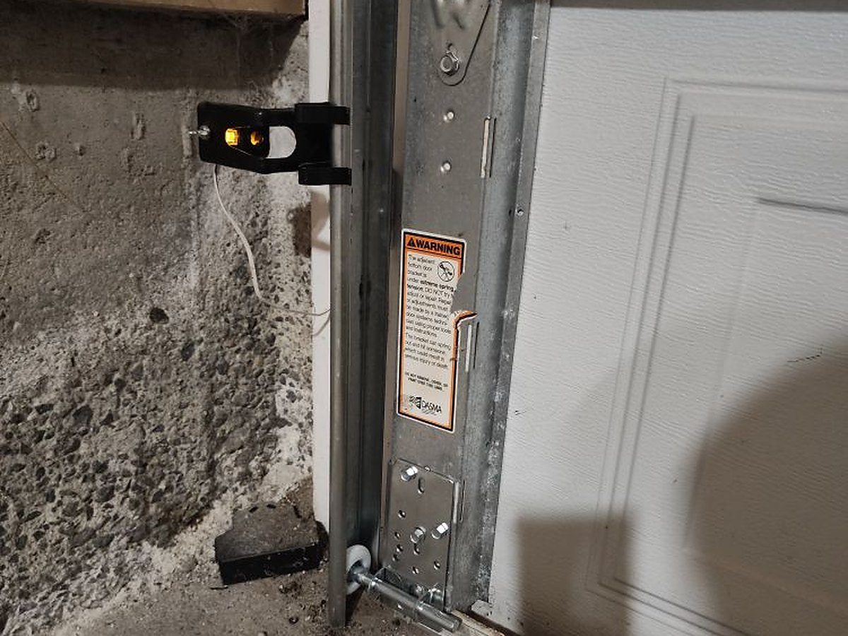 Foot of a vertical garage door track with the roller sitting in its bracket beside the lift cable, and the wall-mounted safety sensor next to the opening